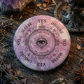 Ceramic pendulum board with all seeing eye design, on a wooden surface with herbs and crystals.