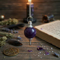Amethyst sphere pendulum hanging over a wooden surface, with a book, feather and token