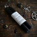 Black spell candle with a label on a wooden surface with dried flowers and a pentagram charm.