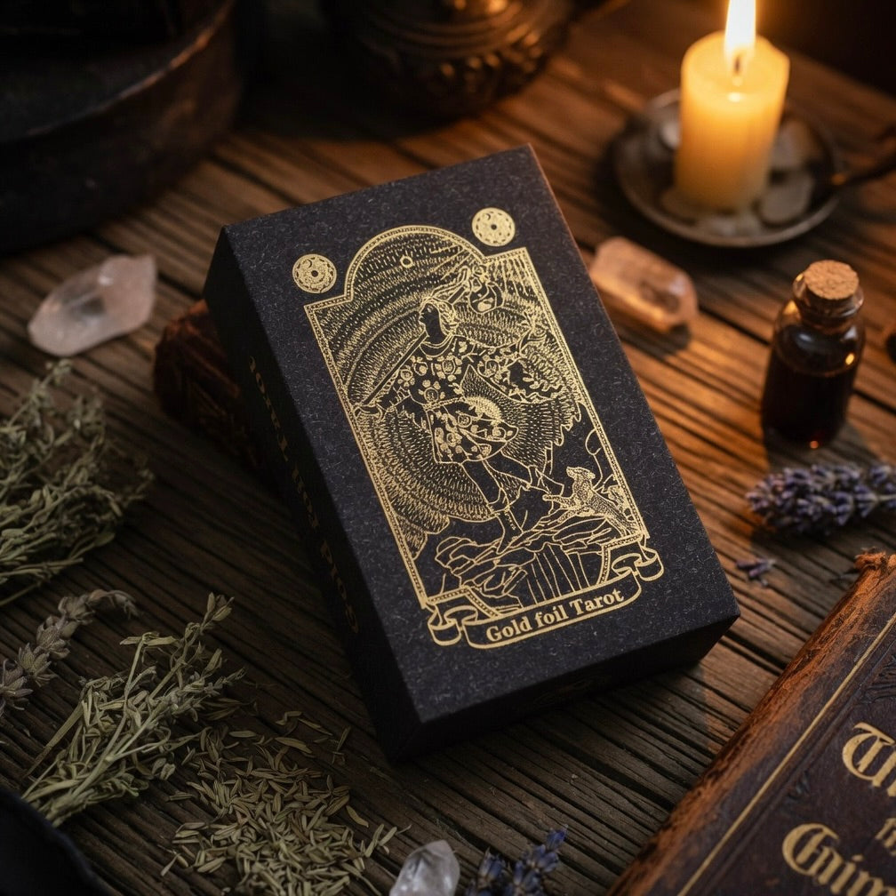 Black and gold foil tarot box on a wooden surface with candle, book and herbs
