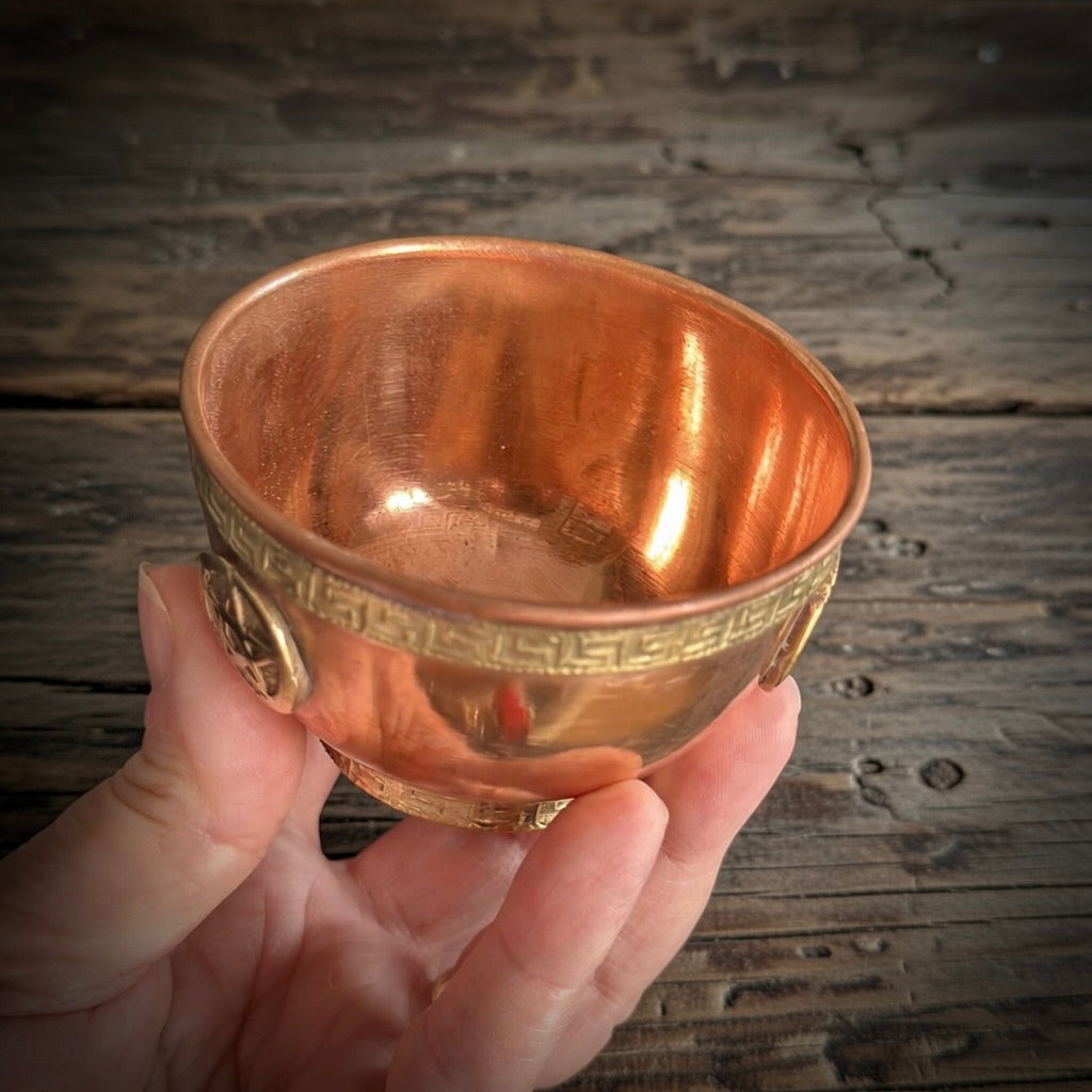 Hand holding a copper altar bowl with intricate designs against a wooden background