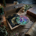 Decorative pendulum divination set with moonlit shadow design on a wooden box surrounded by herbs and books on a rustic surface.