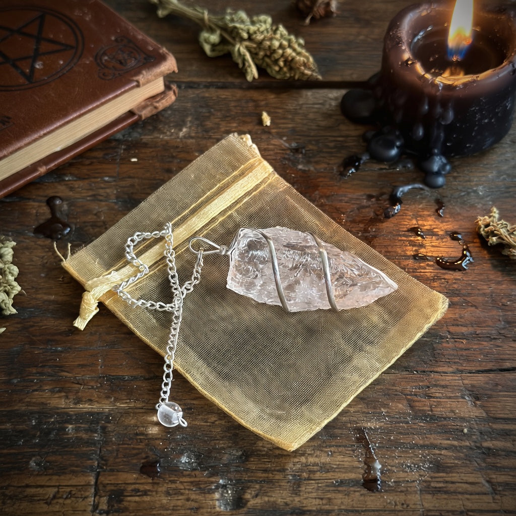 Raw rock quartz pendulum and organza pouch on a wooden surface with a candle and herbs.