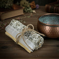 Bundled selenite, sage and palo santo sticks with a pentagram symbol next to a copper smudge bowl on a wooden surface.
