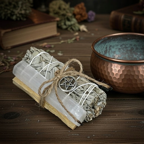 Bundled selenite, sage and palo santo sticks with a pentagram symbol next to a copper smudge bowl on a wooden surface.