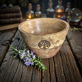 Wooden bowl with tree of life design on a wooden surface with rosemary and witchcraft items in the background