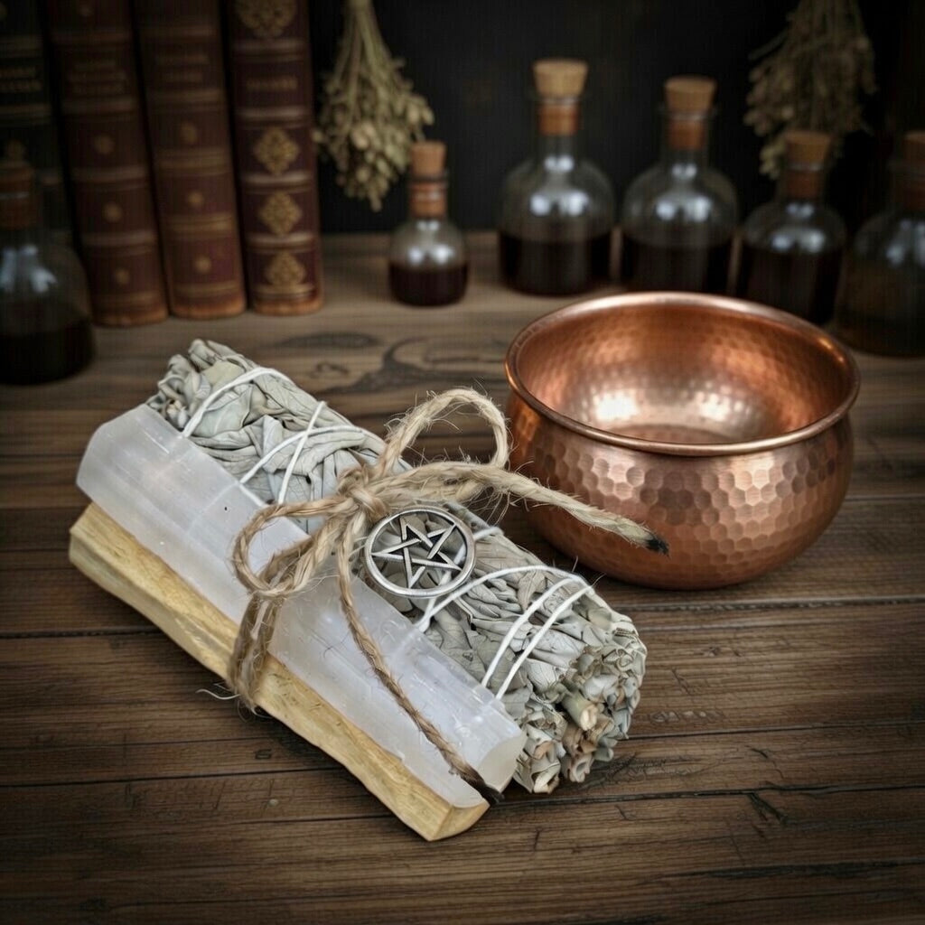 Bundled selenite, sage and palo santo sticks with a pentagram symbol next to a copper smudge bowl on a wooden surface.