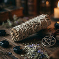 White sage Smudge stick, with lavender, black stones, and pentagram pendant on a wooden surface with a blurred candle in the background.
