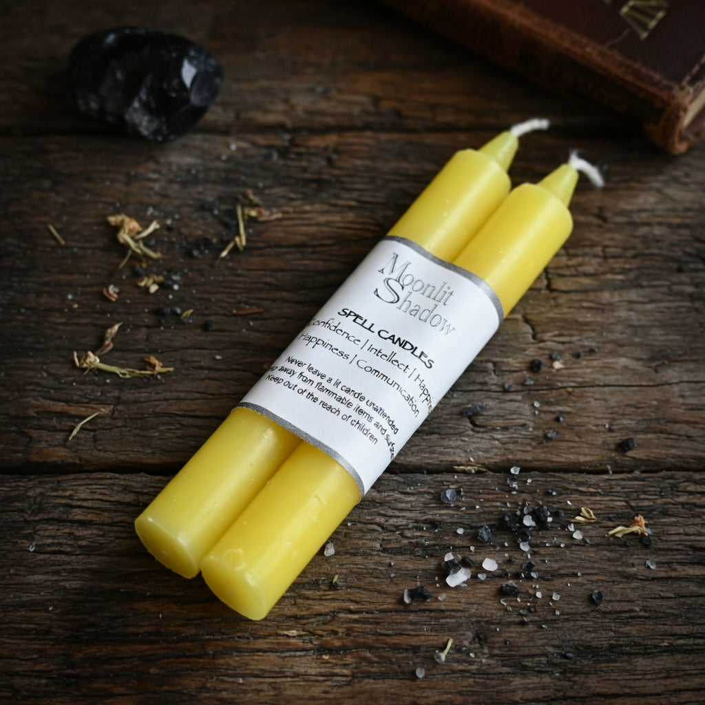 Two yellow spell candles with a label on a wooden surface with herbs and crystals.
